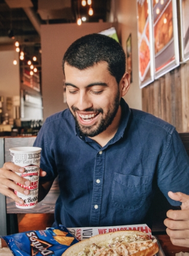 Man wearing blue shirt holding a drink and looking at his sandwich and chips